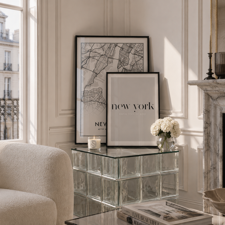 Two framed minimalist art prints with black frames are displayed on a glass block side table in a bright, elegant room. One print features a black-and-white street map of New York, and the other has the text 'new york' in black.