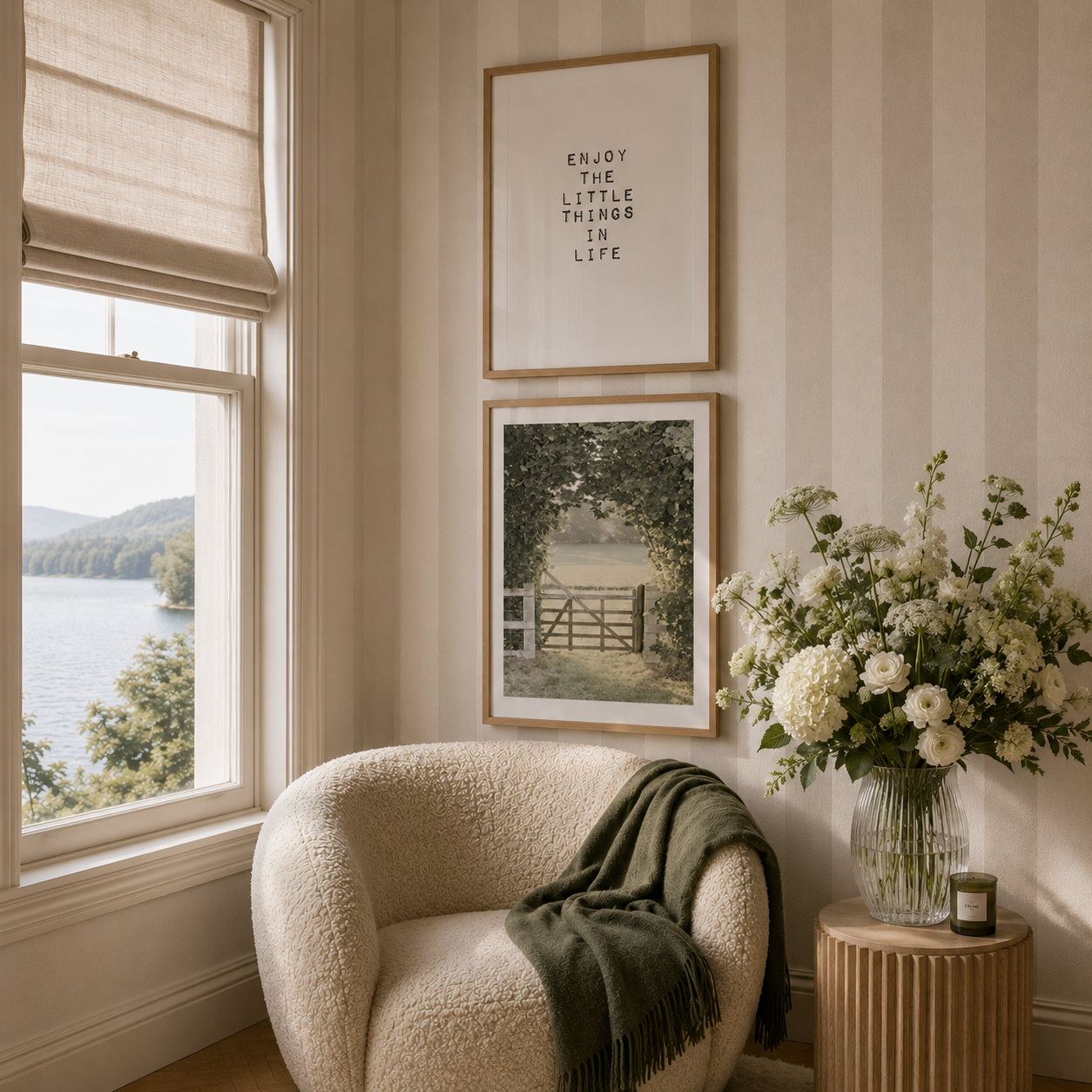 A styled interior corner with a cream boucle armchair, a dark green throw blanket, a ribbed wood side table holding a glass vase of white flowers, and two framed wall prints above a window overlooking water.