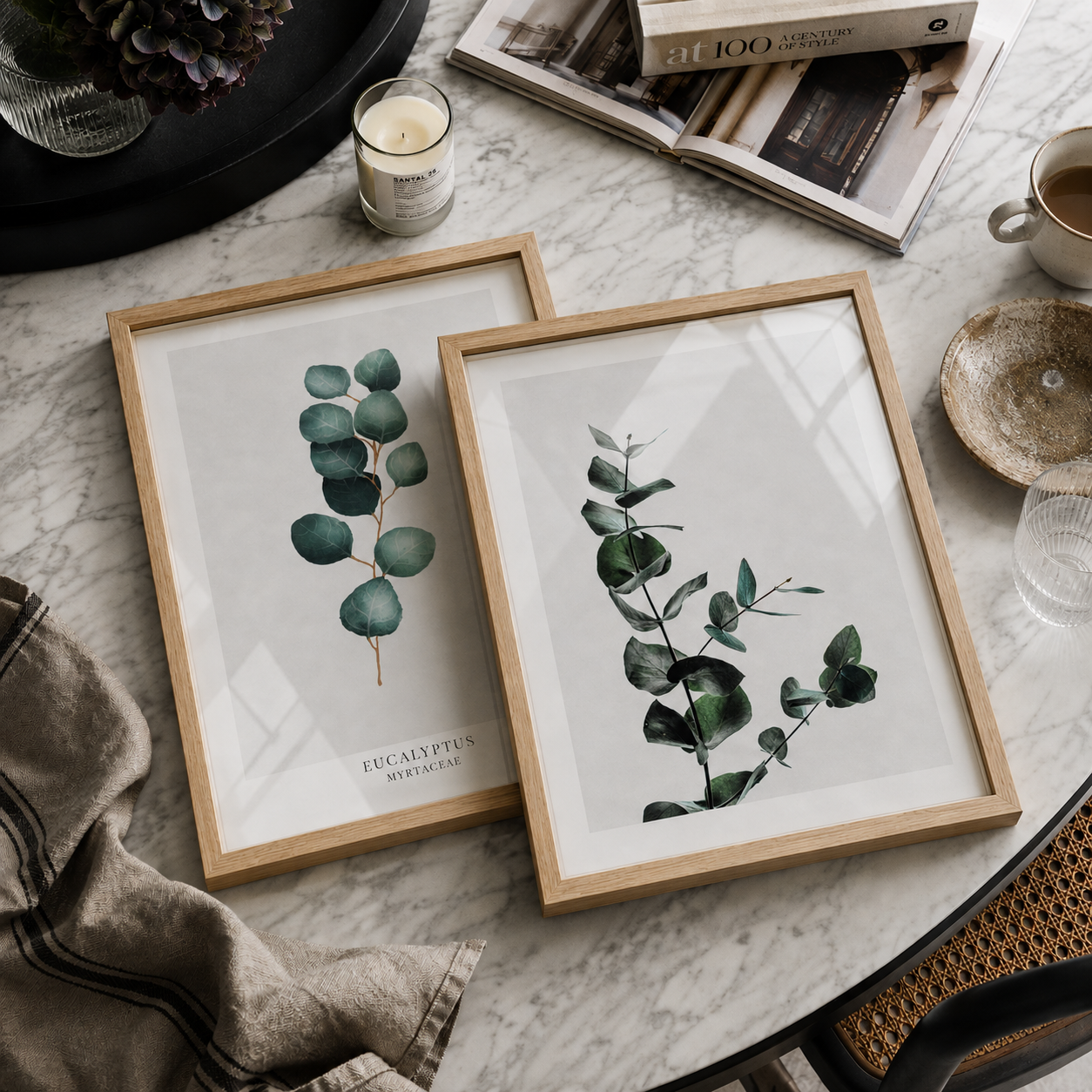Two framed botanical prints featuring eucalyptus stems, styled on a marble tabletop with books, a candle, and a cup nearby.