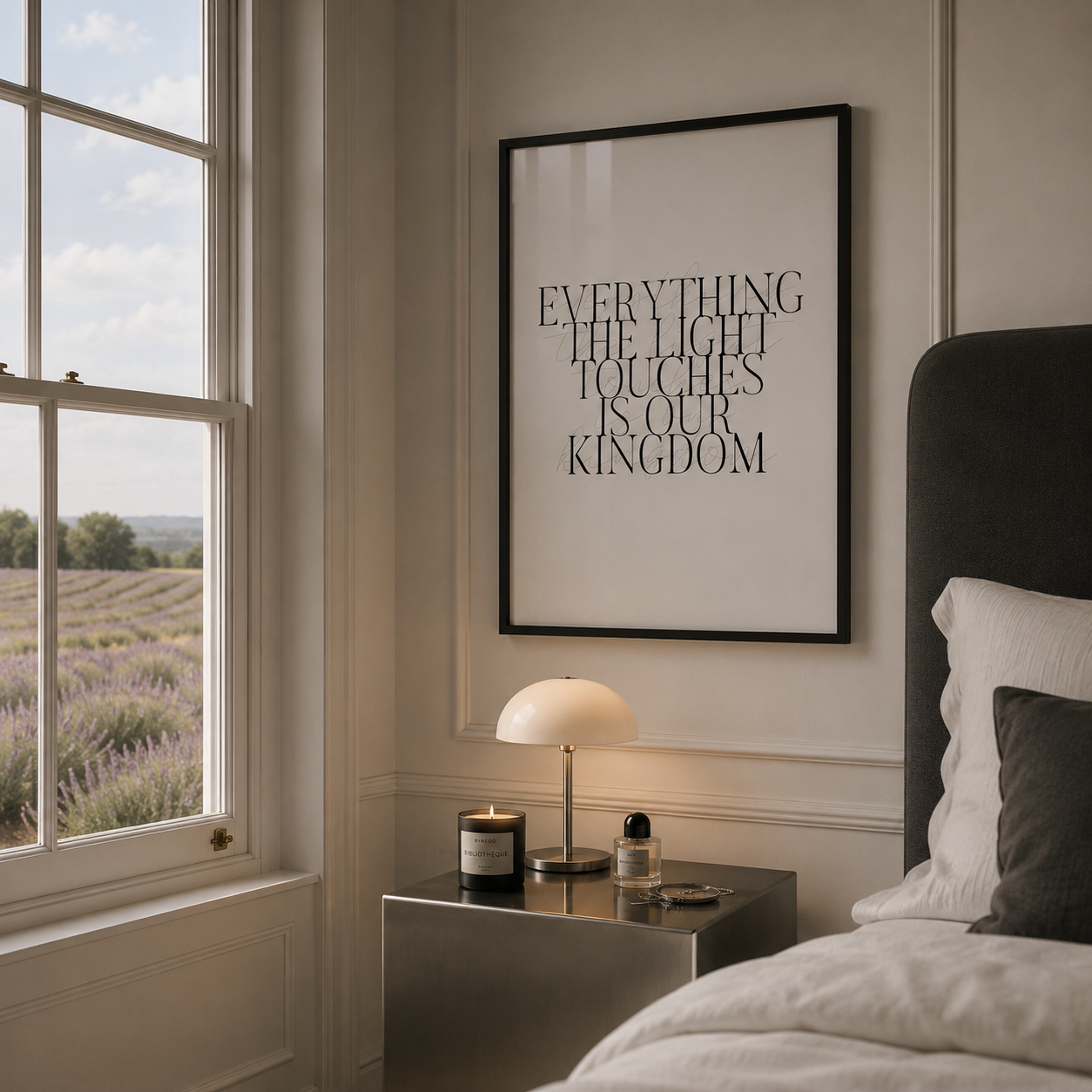 A framed typographic wall art print hangs on a beige bedroom wall above a nightstand, with a window, lamp, and bed visible in the room. The print reads, 'EVERYTHING THE LIGHT TOUCHES IS OUR KINGDOM.