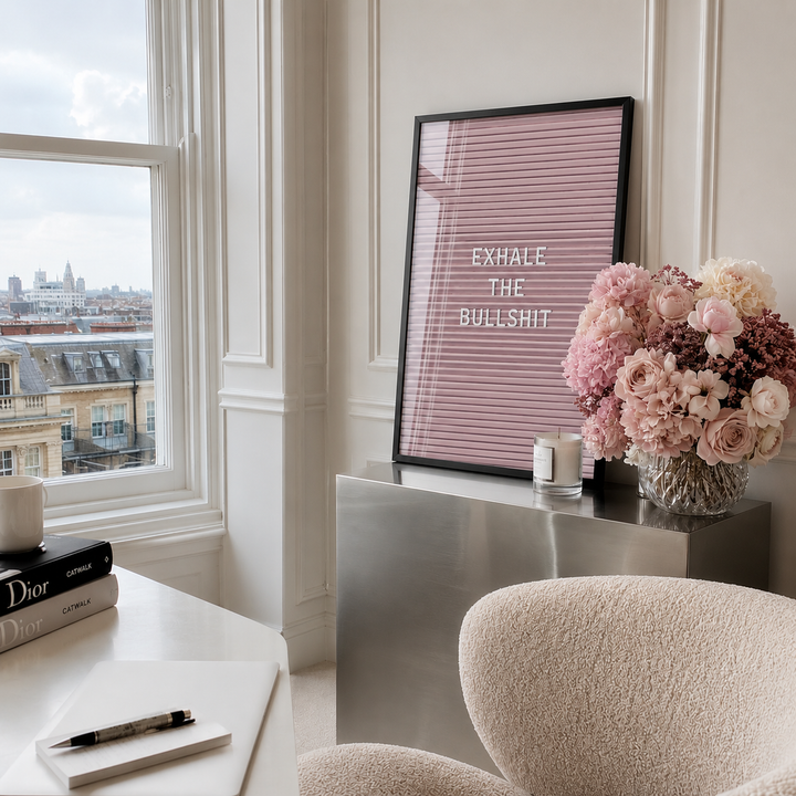 A framed pink striped art print with the text 'EXHALE THE BULLSHIT' in white, styled in a bright, modern room next to a window with a view of a city skyline. The print is displayed on a metallic sideboard next to a vase of pink and white flowers.