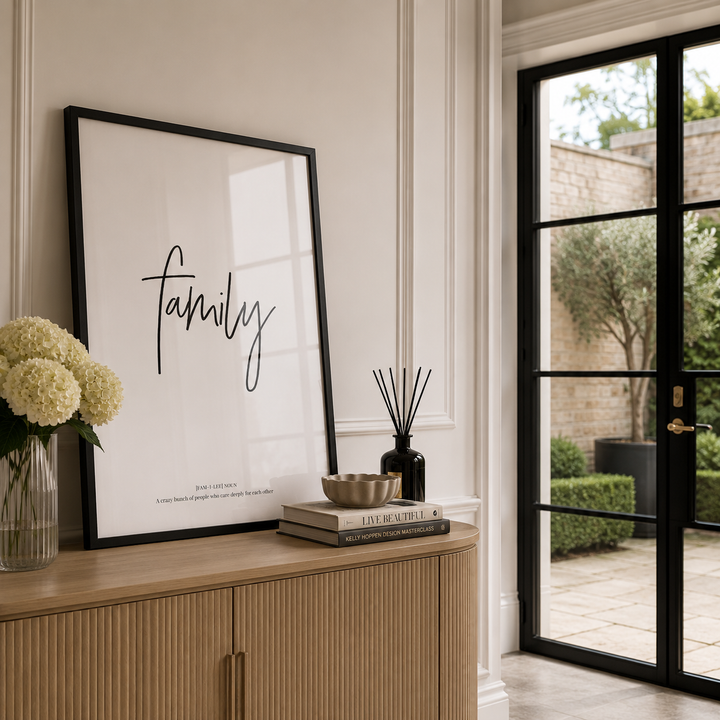 A framed black-and-white art print with the word 'family' in script, leaning on a light wood console beside a vase of white flowers and a reed diffuser, with a black-framed glass door opening to a garden in the background.
