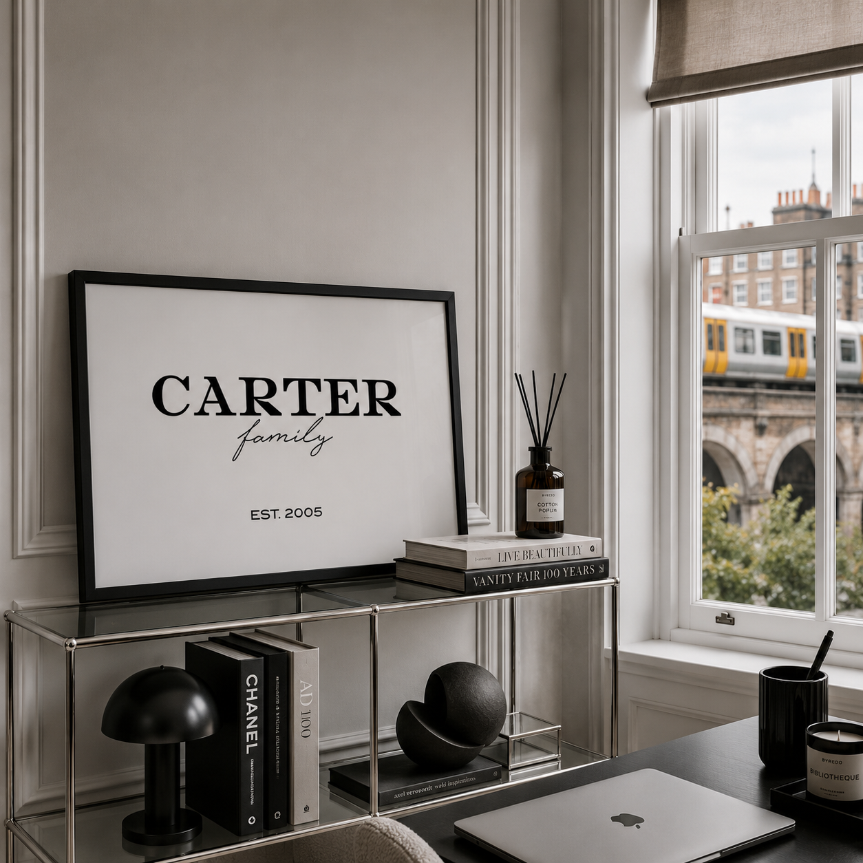 A framed black-and-white typographic print reading 'CARTER family EST. 2005' displayed on a glass shelf in a styled home office setting next to a window with a train visible outside.