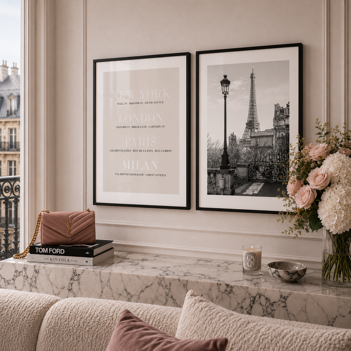 Two framed wall art prints hang above a marble console table in a styled interior. The left print is a minimalist city names poster, and the right print is a black-and-white Paris scene with the Eiffel Tower.
