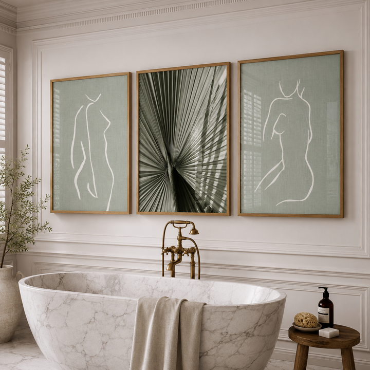 Three framed wall art prints hang above a freestanding marble bathtub in a styled bathroom. The left and right frames show minimalist white line drawings of female figures on a muted sage green background, and the center frame shows a dark green palm frond pattern.