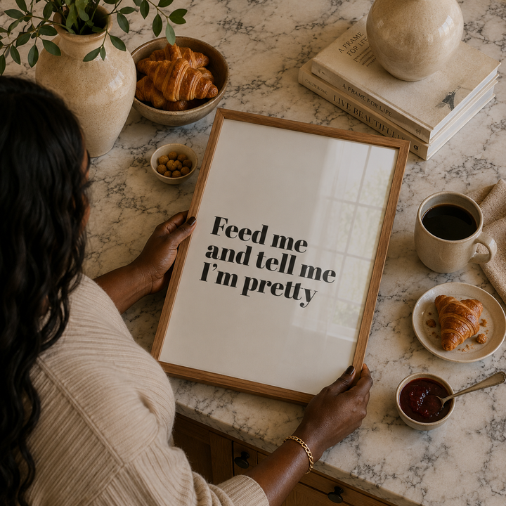 Woman holding a framed poster with text "Feed me and tell me I'm pretty" in luxury kitchen.