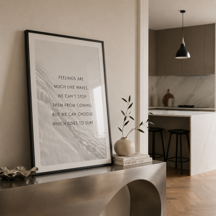 A framed wall art print with a quote about feelings and waves, leaning on a metallic console table in a modern interior. The print has a black frame, white mat, and black text on a light gray marbled background.