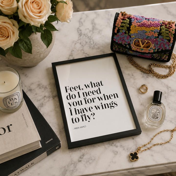 A styled flat-lay featuring a framed black-and-white quote print with the text 'Feet, what do I need you for when I have wings to fly?' attributed to Frida Kahlo. The print is arranged on a white marble surface alongside a multicolor Valentino beaded clutch bag, a candle, books, a perfume bottle, rings, and a gold necklace.