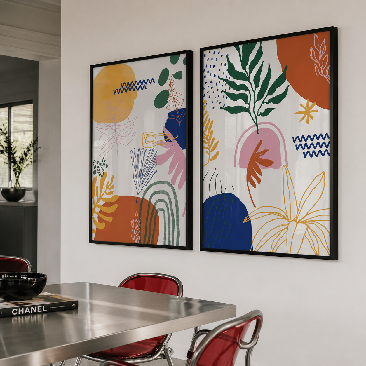 Two framed abstract botanical wall art prints hang on a white wall above a stainless steel dining table with red chairs in a modern interior setting.