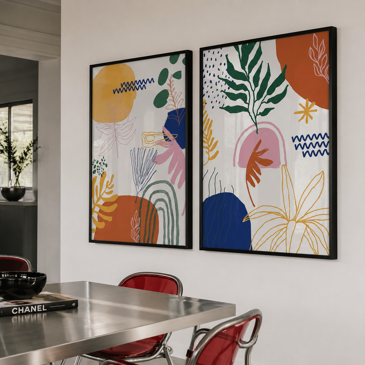 Two framed abstract botanical wall art prints hang on a white wall above a stainless steel dining table with red chairs in a modern interior setting.
