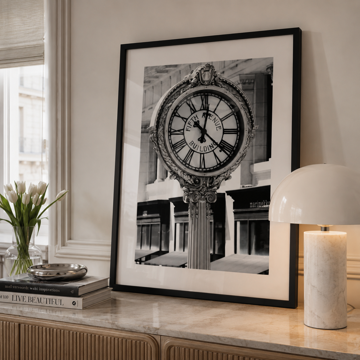 A large black and white framed art print of a vintage street clock stands on a marble console table in a styled room setting, next to a vase of tulips and a lit table lamp.