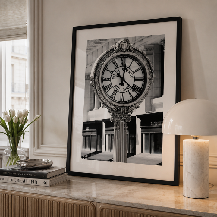 A large black and white framed art print of a vintage street clock stands on a marble console table in a styled room setting, next to a vase of tulips and a lit table lamp.