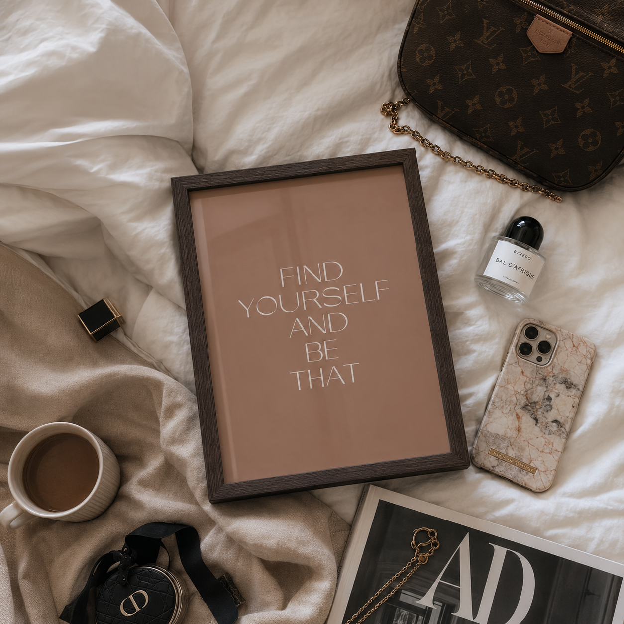 A framed motivational art print with the text 'FIND YOURSELF AND BE THAT' in white on a warm beige background, styled in a flat lay on white bedding with a brown monogram handbag, a coffee cup, a marble-patterned phone case, and other lifestyle accessories.
