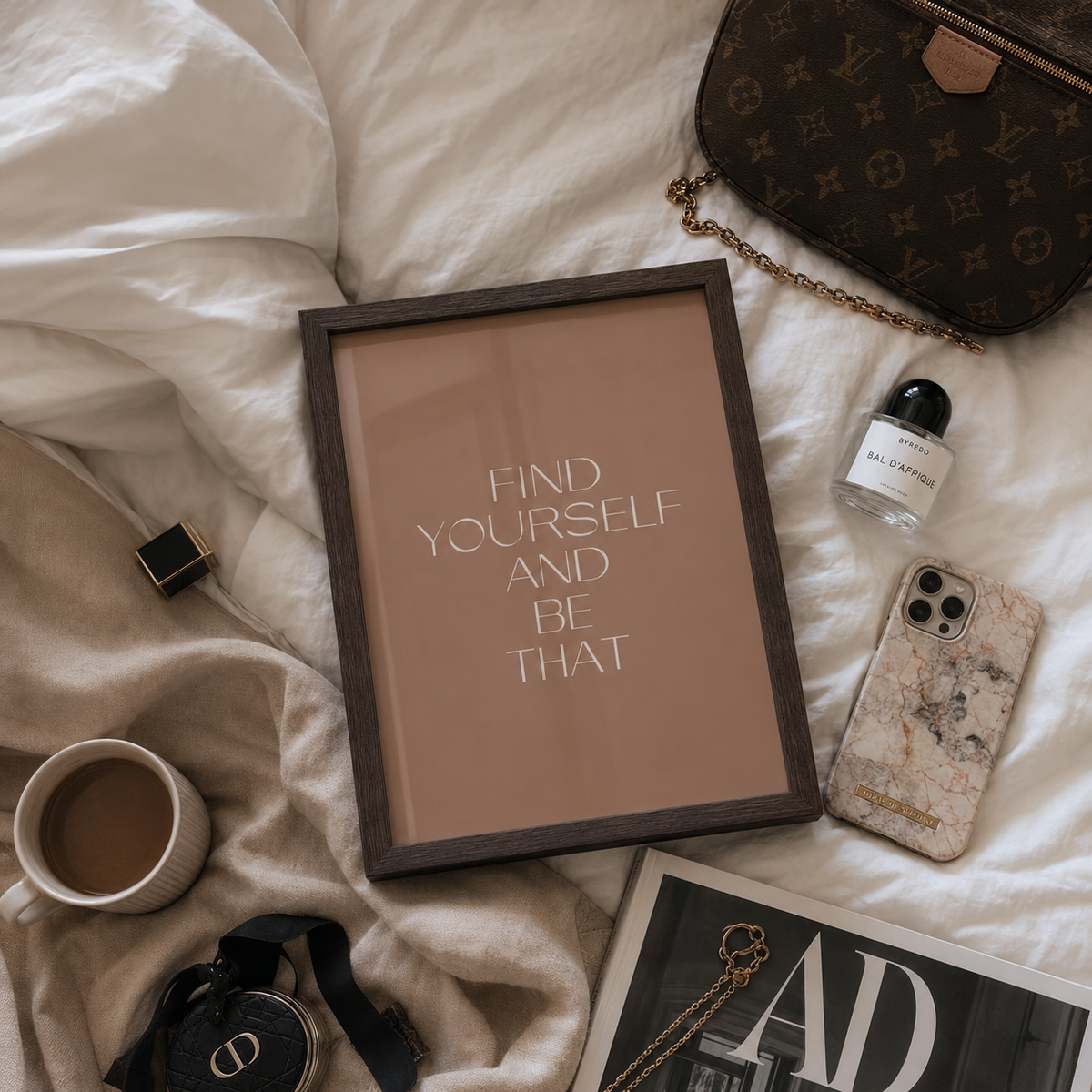 A framed motivational art print with the text 'FIND YOURSELF AND BE THAT' in white on a warm beige background, styled in a flat lay on white bedding with a brown monogram handbag, a coffee cup, a marble-patterned phone case, and other lifestyle accessories.