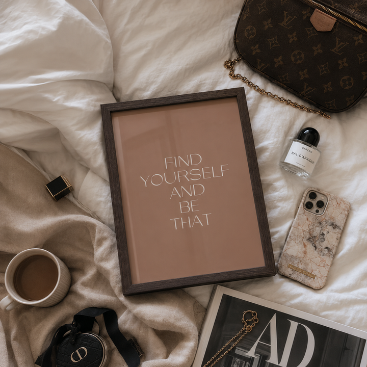 A framed motivational art print with the text 'FIND YOURSELF AND BE THAT' in white on a warm beige background, styled in a flat lay on white bedding with a brown monogram handbag, a coffee cup, a marble-patterned phone case, and other lifestyle accessories.