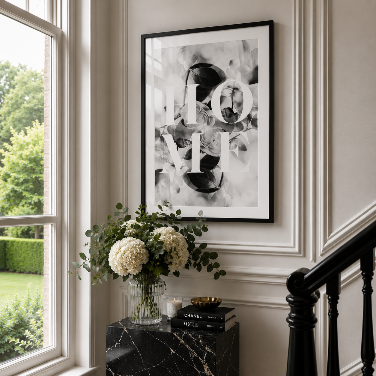 A framed black and white abstract art print with the word 'HOME' in large white letters, hanging on a beige wall in a styled interior next to a window and a black marble side table.
