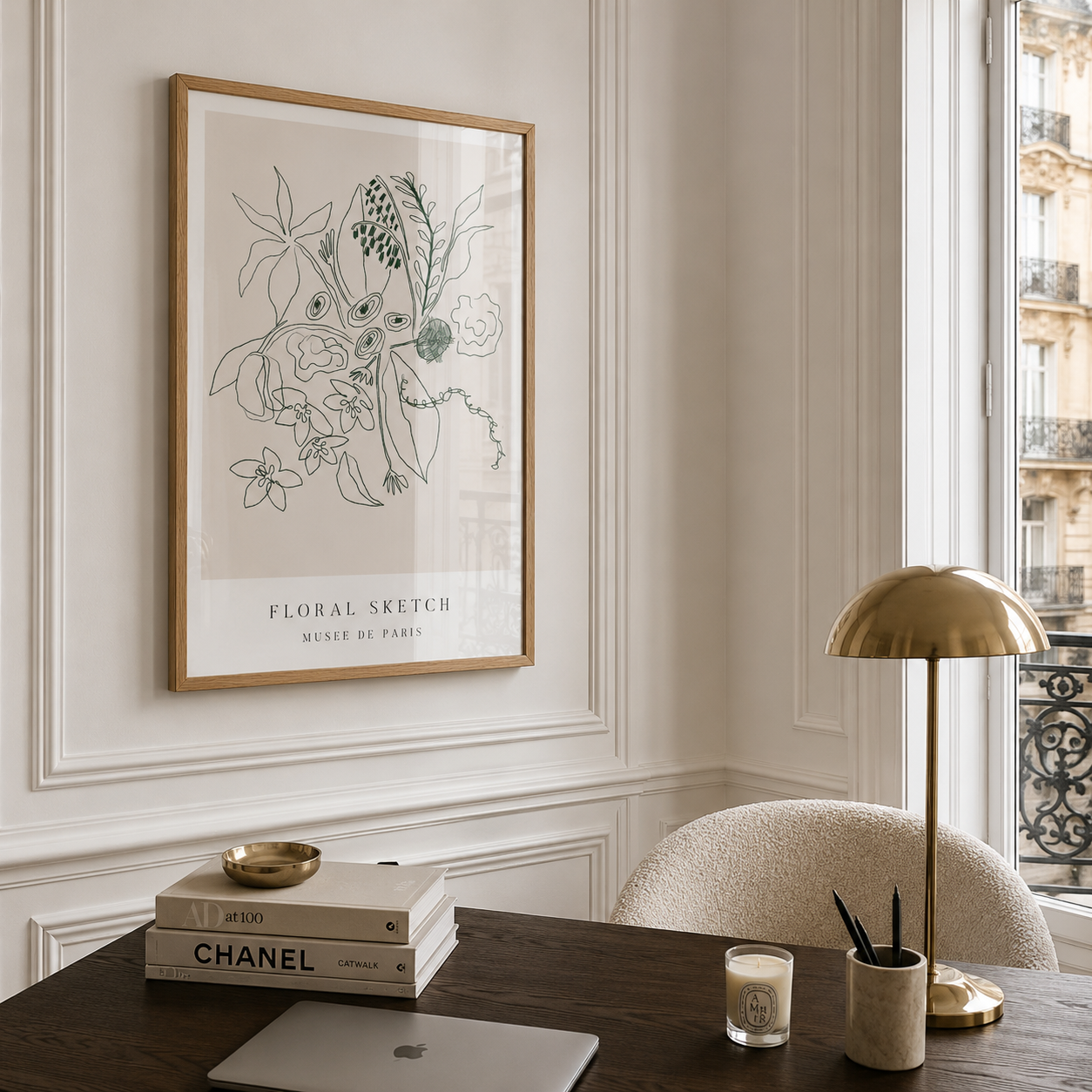 A framed botanical art print with black line drawings and the text 'FLORAL SKETCH / MUSE DE PARIS' hangs on a cream paneled wall above a dark wood desk with a brass lamp, books, and a laptop in a bright interior setting.
