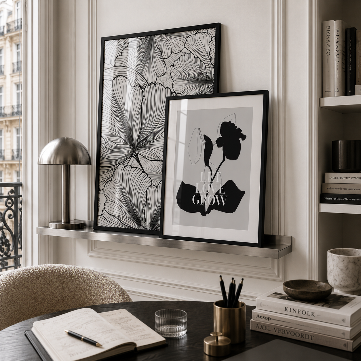Two framed black and white botanical art prints displayed on a silver floating shelf in a styled home office setting with a desk, lamp, and books.