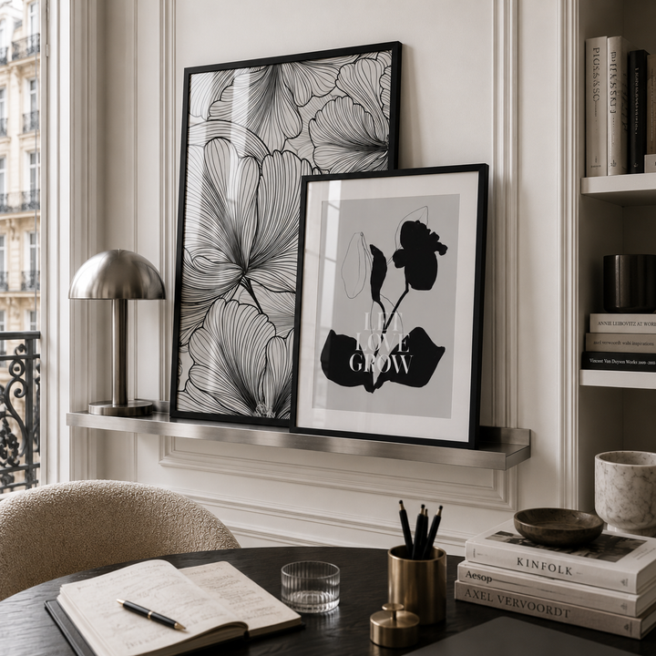 Two framed black and white botanical art prints displayed on a silver floating shelf in a styled home office setting with a desk, lamp, and books.
