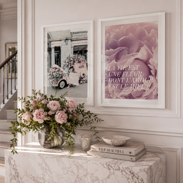A styled interior scene with two framed floral wall art prints hanging above a marble console table. The right print features large pink flowers and French text reading 'LA VIE EST UNE FLEUR DONT L'AMOUR EST LE MIEL.