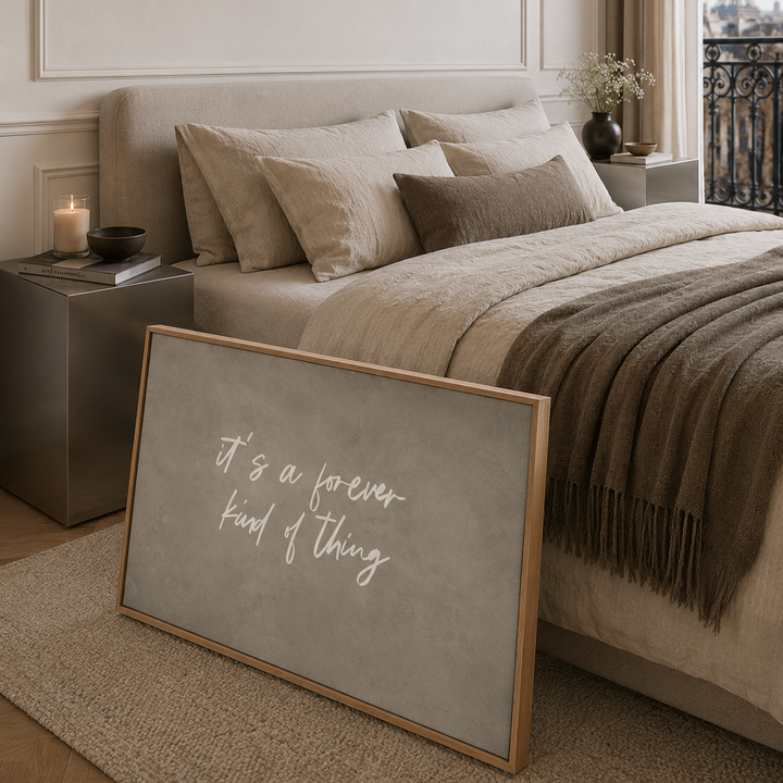 A framed large canvas art print with the quote 'it's a forever kind of thing' in white script on a textured neutral background, styled in a parisian bedroom next to a bed with neutral beige bedding.
