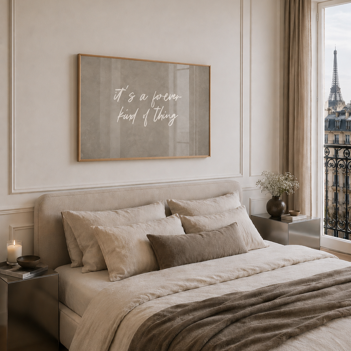 A styled bedroom interior with a bed dressed in beige and taupe bedding, a large oak framed wall art print above the headboard reading 'it's a nice kind of thing', and a window showing the Eiffel Tower outside.