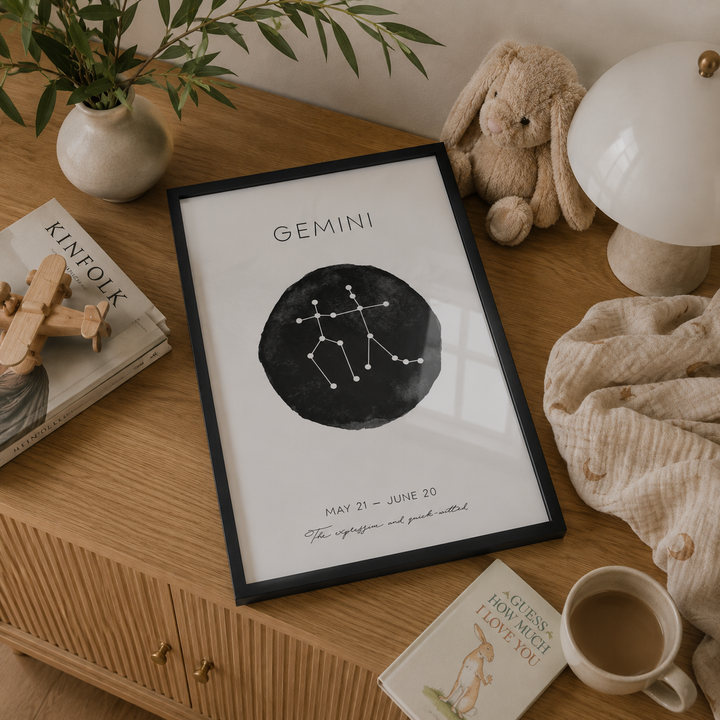 A framed Gemini zodiac art print displayed on a wooden dresser in a cozy room setting, surrounded by a plush bunny, a lamp, a cup of tea, and other decorative items.