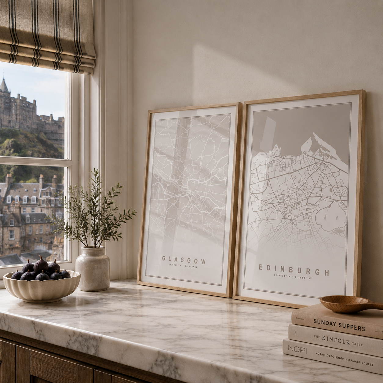 Two framed minimalist city map prints leaning on a marble countertop in a bright kitchen. The left print is labeled Glasgow and the right print is labeled Edinburgh.