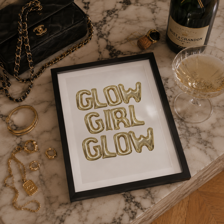 A framed art print featuring the phrase 'GLOW GIRL GLOW' in raised gold letters on a white background, enclosed in a black frame. The frame is styled on a white marble surface alongside a black quilted handbag, gold jewelry, a champagne bottle, and a champagne flute.