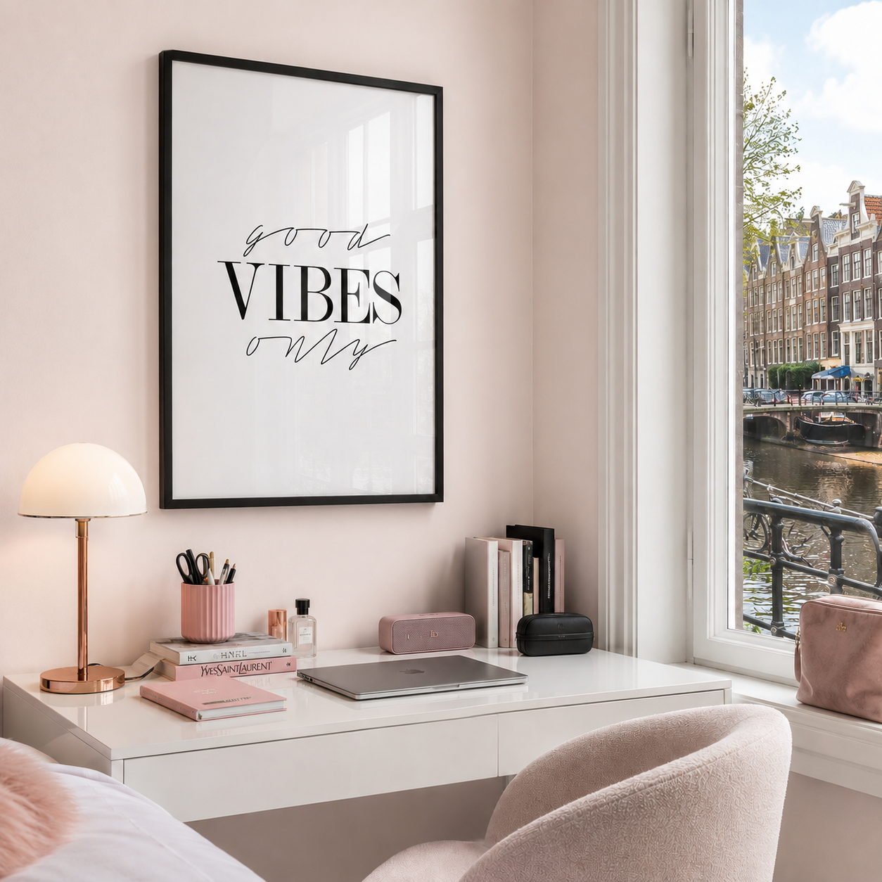 A framed wall art print with the words 'good VIBES only' in black script and serif lettering on a white background, hanging above a white desk in a softly styled room with pink walls, a lamp, books, and a window overlooking an Amsterdam canal.