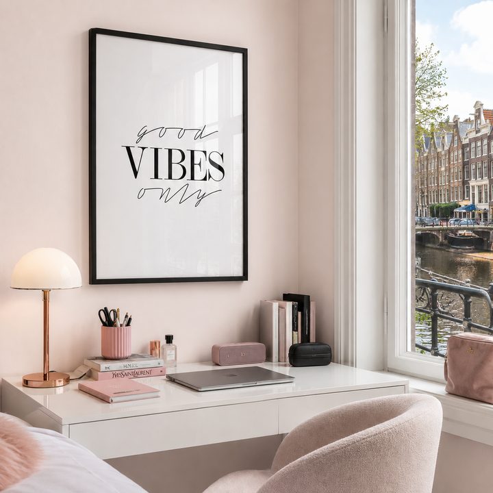 A framed wall art print with the words 'good VIBES only' in black script and serif lettering on a white background, hanging above a white desk in a softly styled room with pink walls, a lamp, books, and a window overlooking an Amsterdam canal.