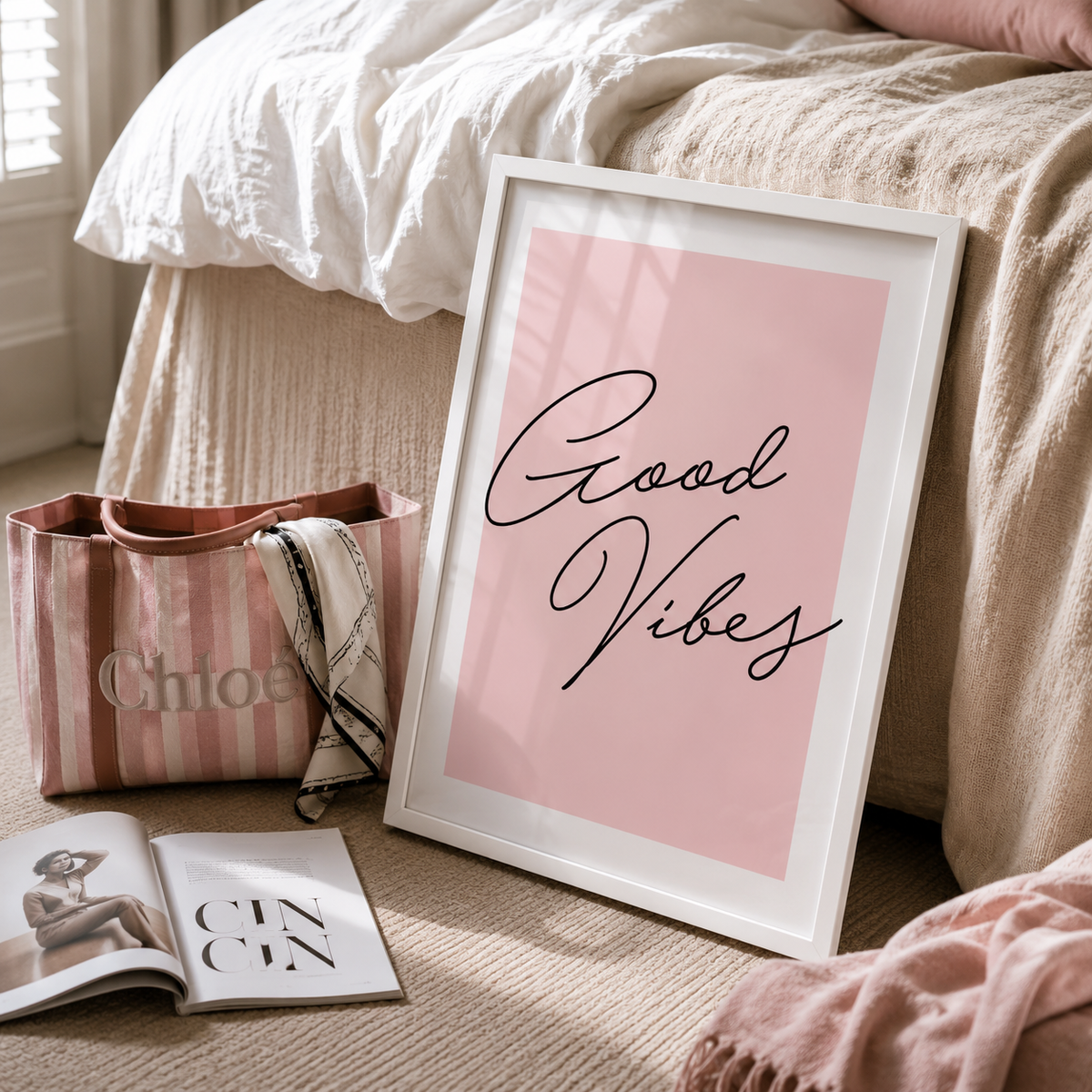 A framed art print with the words 'Good Vibes' in black script on a soft pink background, leaning against a bed in a cozy bedroom setting. The white frame is shown alongside a pink striped tote bag, an open magazine, and a pink blanket.