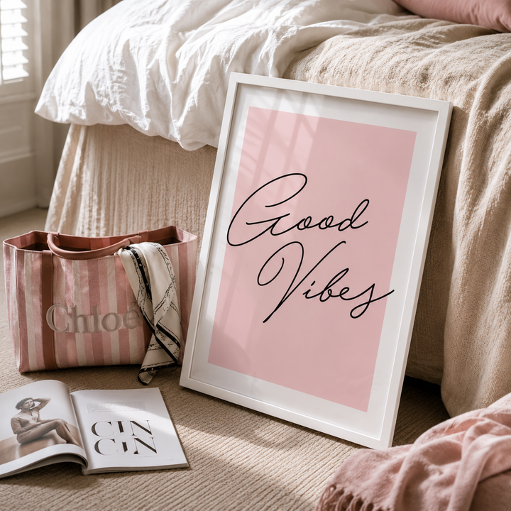 A framed art print with the words 'Good Vibes' in black script on a soft pink background, leaning against a bed in a cozy bedroom setting. The white frame is shown alongside a pink striped tote bag, an open magazine, and a pink blanket.