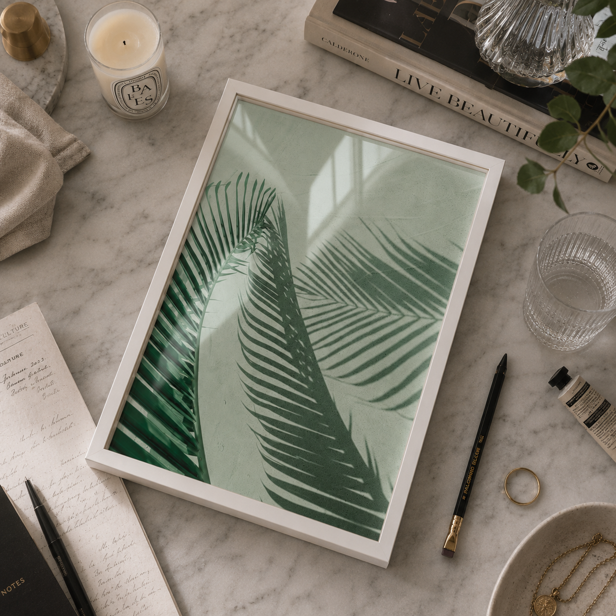 A framed botanical art print featuring green palm leaves on a pale mint background, styled in a flat-lay arrangement on a marble surface with a candle, book, glass, and stationery.