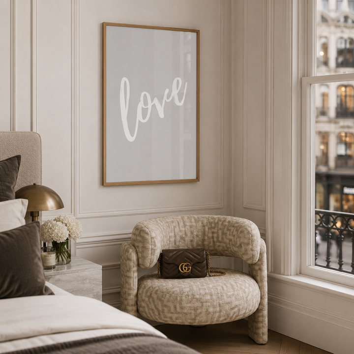 A framed wall art print with the word 'love' in white script on a light gray background, hanging in a styled bedroom corner above a patterned Dunelm beige armchair with a dark brown Gucci handbag resting on it.