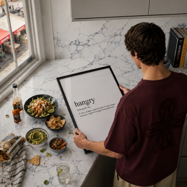 Person holding a framed definition of 'hangry' in a kitchen with food and drinks on the counter.