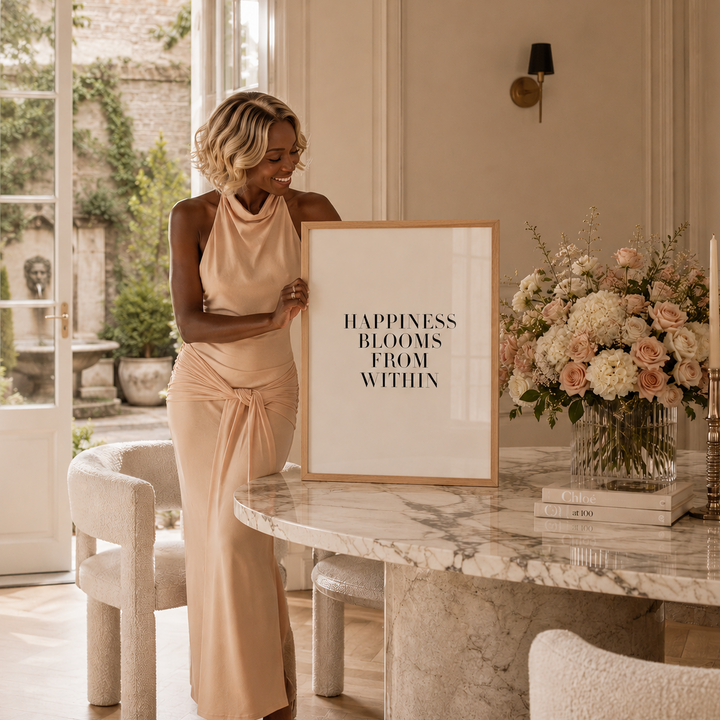 A woman in a beige sleeveless dress stands indoors beside a marble table holding a framed art print that reads 'HAPPINESS BLOOMS FROM WITHIN.' The room has cream walls, large windows, a floral arrangement, and neutral-toned furniture.