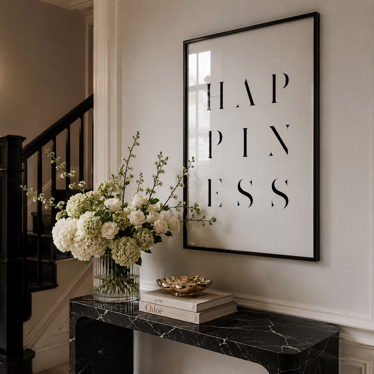 A framed vertical wall art print with the word 'HAPPINESS' in large black serif letters on a white background, hanging above a black marble console table with a floral arrangement and books in a styled interior.
