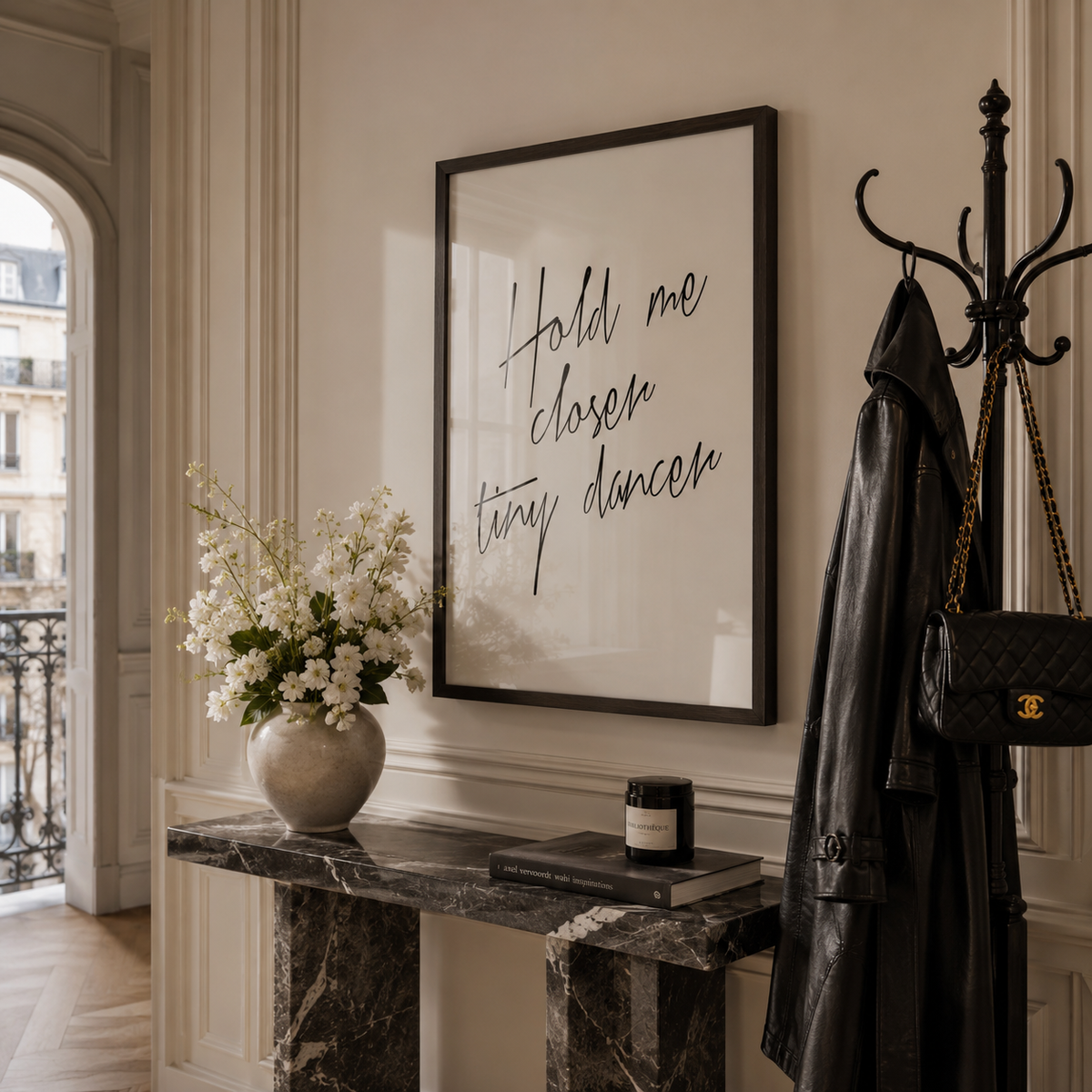 A framed wall art print with the phrase 'Hold me closer tiny dancer' in black script on a white background, hanging in a styled interior above a marble console table with flowers, a candle, and a black coat on a coat rack.