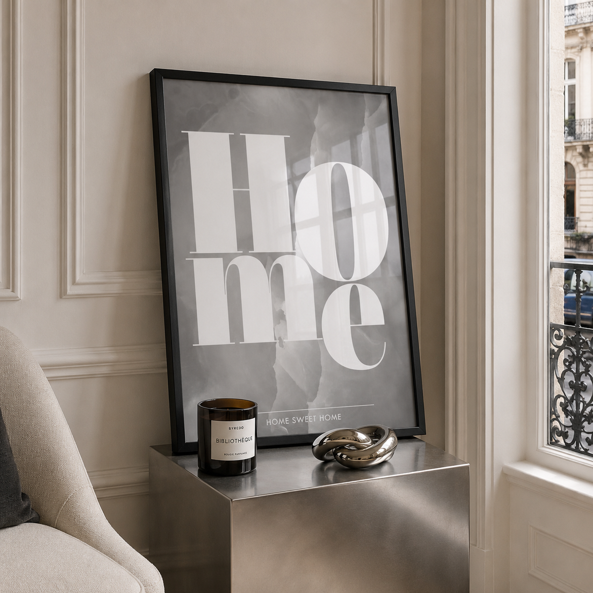 A framed typographic wall art print leaning on a metallic side table in a styled interior. The print features the word 'home' in large white letters over a smoky gray background, with smaller text reading 'home sweet home' near the bottom.