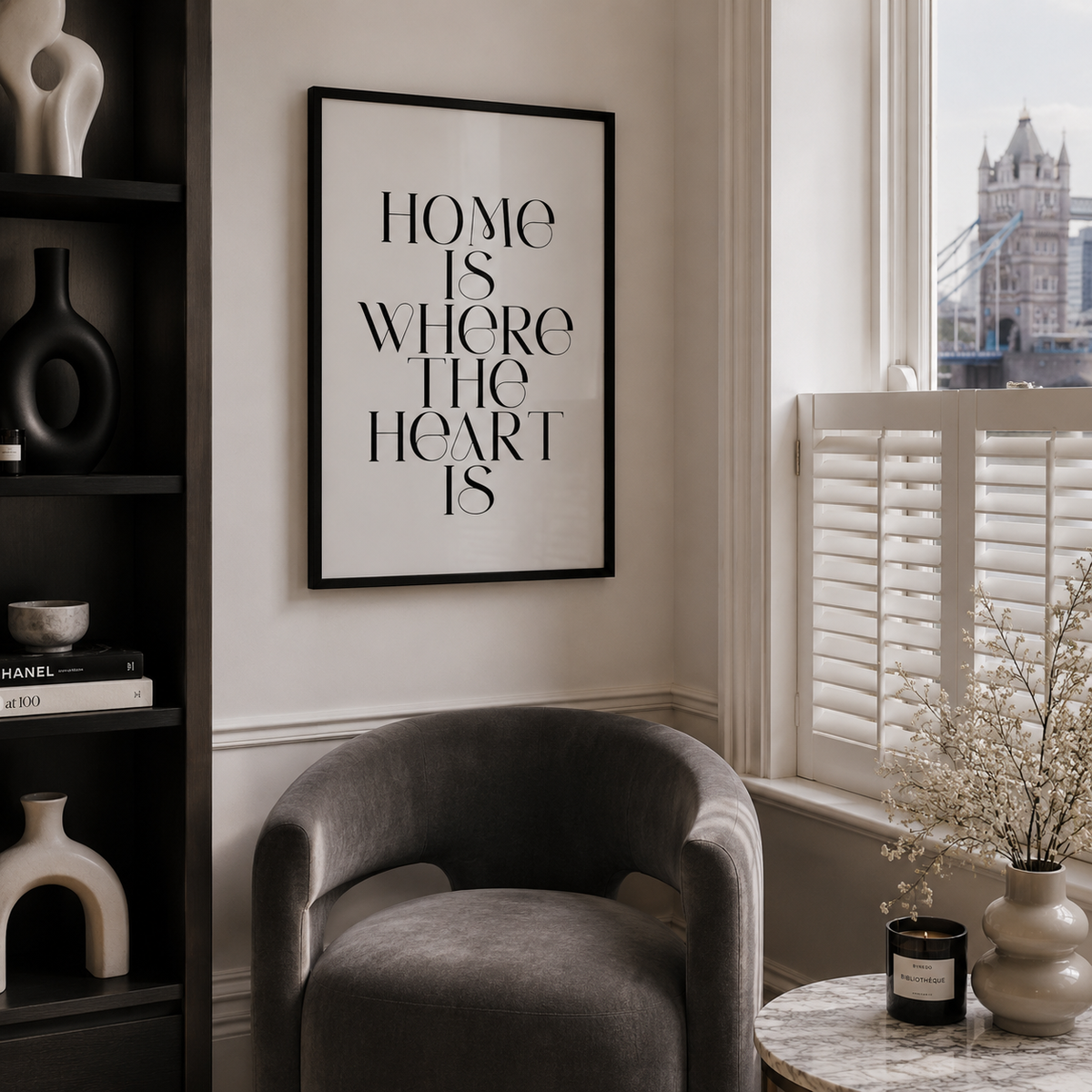 A framed black and white typographic wall art print reading 'HOME IS WHERE THE HEART IS' hangs on a cream wall in a styled living room with a gray armchair, dark shelving, and a window with shutters.