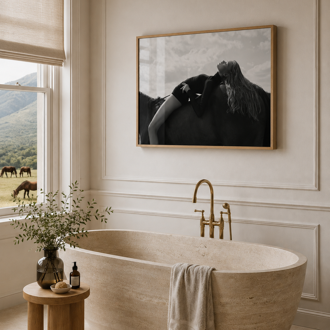 A framed black and white art print depicting a woman kissing a horse, hung on a beige wall above a freestanding stone bathtub in a bright, elegant bathroom.