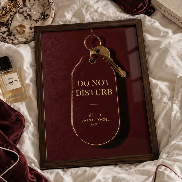A dark burgundy hotel key tag with gold lettering reading 'DO NOT DISTURB' and 'HÔTEL SAINT ROCHE PARIS', attached to a gold key ring with a brass key, shown against a matching burgundy background.