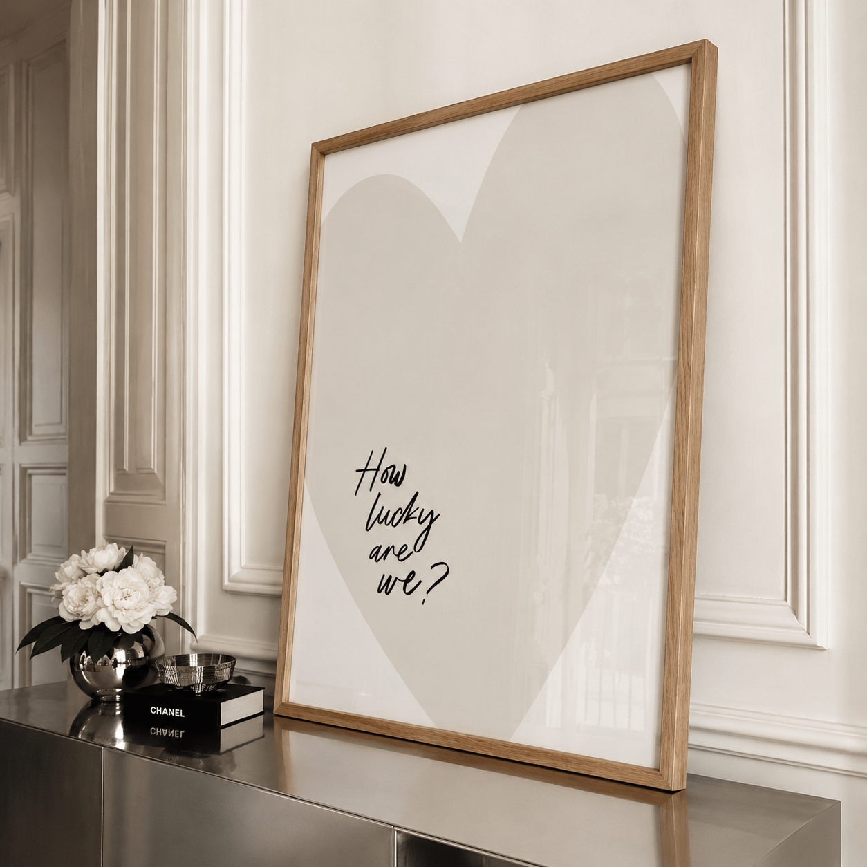 A framed art print with a light wood frame leans against a white paneled wall on a metallic console table. The print features a large beige heart shape and the handwritten text 'How lucky are we?' in black.