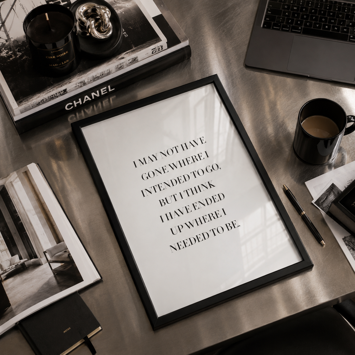 A framed typographic wall art print with black text on a white background, styled on a metallic desk with a laptop, coffee cup, notebook, and other office props.