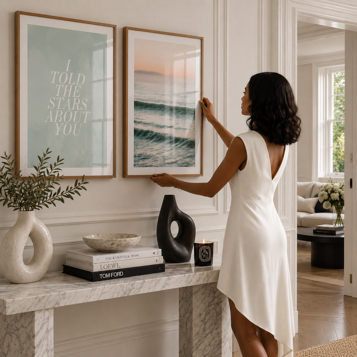 A woman in a white dress hangs a framed ocean print on a white paneled wall in a bright living room. Another framed art print with text hangs beside it, and a marble console table with decorative objects is below.