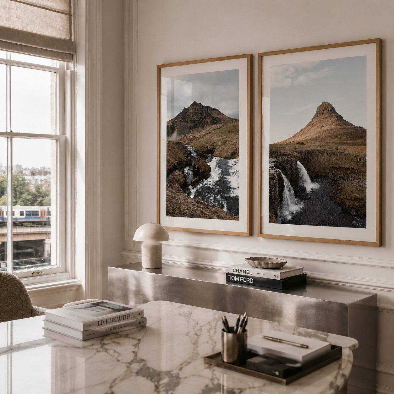 Two framed landscape art prints depicting mountain and waterfall scenes, displayed side-by-side on a beige wall above a marble desk in a modern, sunlit room.