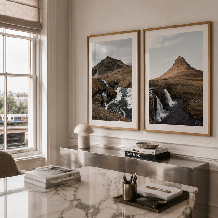Two framed landscape art prints depicting mountain and waterfall scenes, displayed side-by-side on a beige wall above a marble desk in a modern, sunlit room.