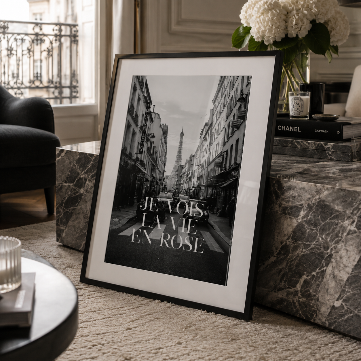 A framed black and white art print leaning against a marble side table in a stylishly decorated room. The print features a Parisian street scene with the Eiffel Tower in the background and the French text 'JE VOIS, LA VIE, EN-ROSE'.