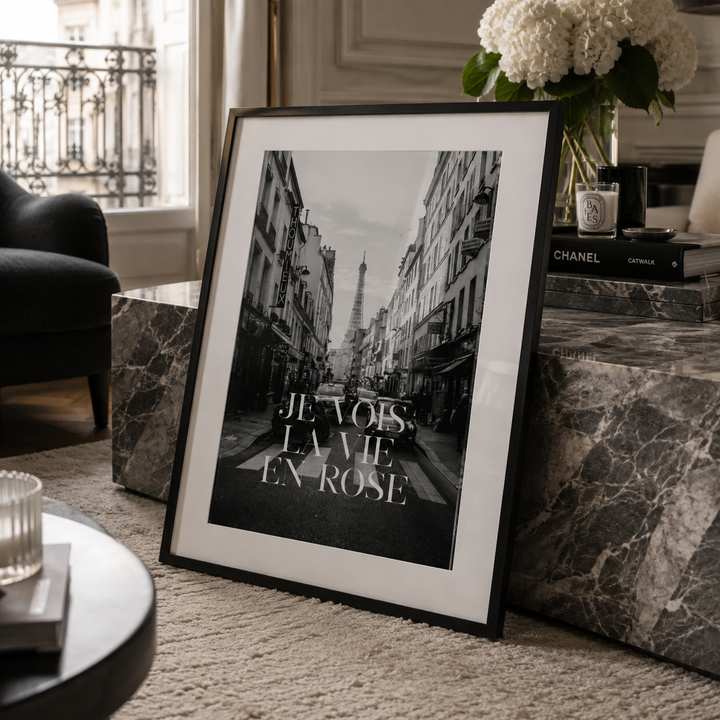 A framed black and white art print leaning against a marble side table in a stylishly decorated room. The print features a Parisian street scene with the Eiffel Tower in the background and the French text 'JE VOIS, LA VIE, EN-ROSE'.